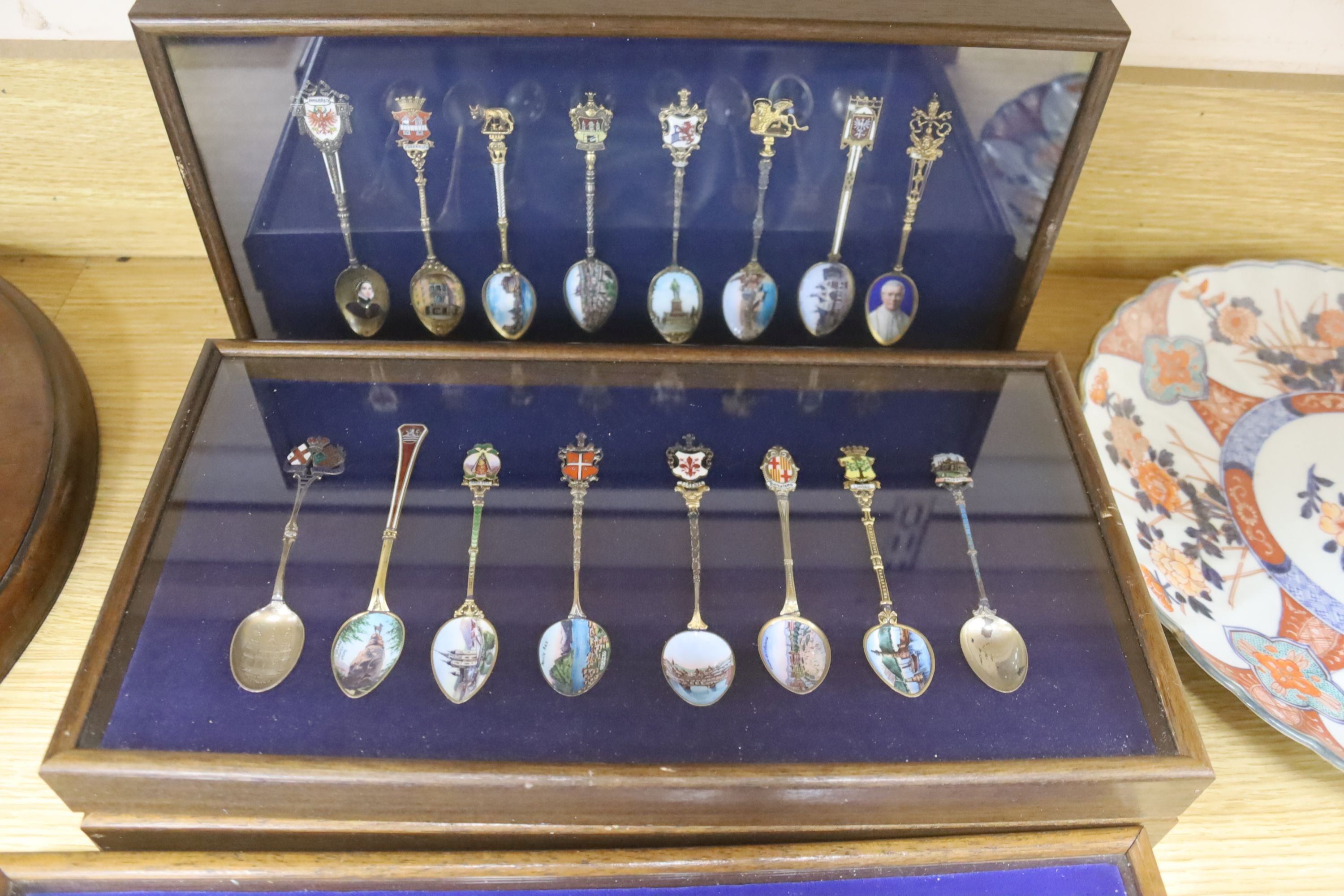 A collection of sixty four 800 standard silver (many gilt) and enamel souvenir spoons, early 20th century, in eight display cases, Including views of London, Germany, Italy, Austria, Spain etc.